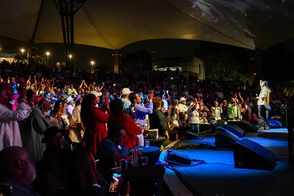 A packed amphitheatre crowd with hands in the air facing the stage at night